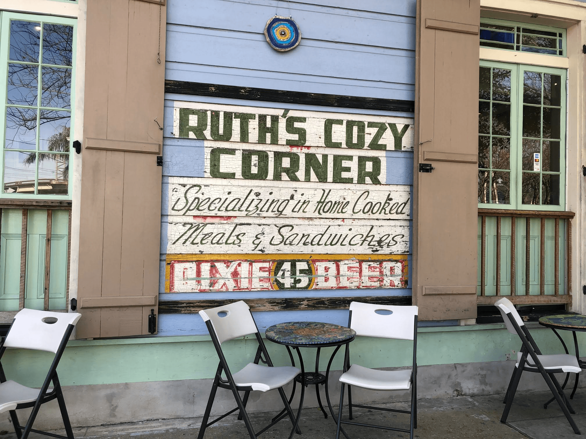 Sign for "Ruth's Cozy Corner" featuring home-cooked meals and sandwiches, with empty chairs outside.