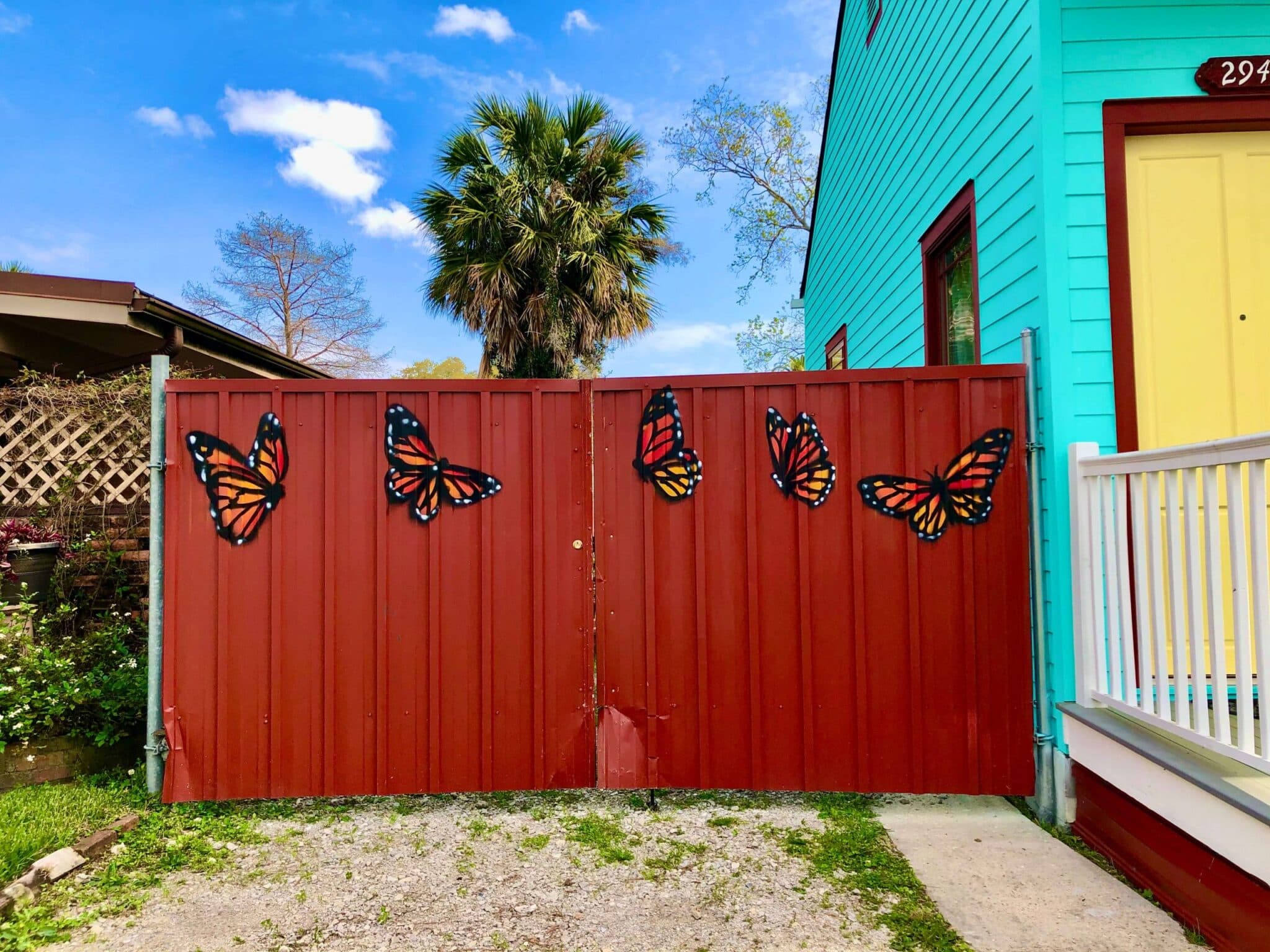 A red metal gate adorned with painted butterflies, set against a colorful house and palm tree backdrop. A red metal gate adorned with painted butterflies, set against a colorful house and palm tree backdrop.