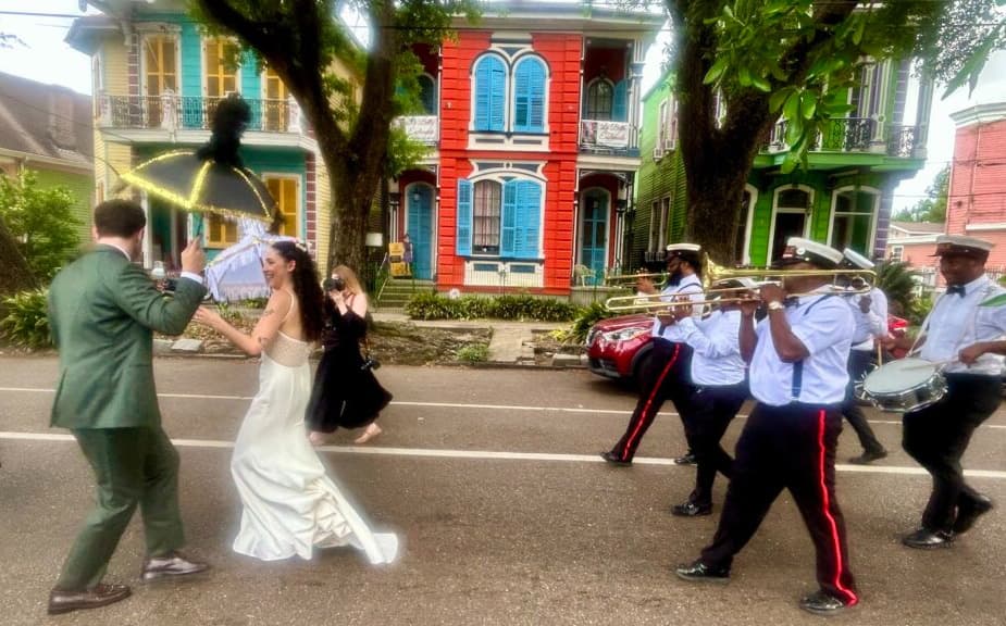 A bride and groom dance down the street while a brass band plays in a vibrant neighborhood.